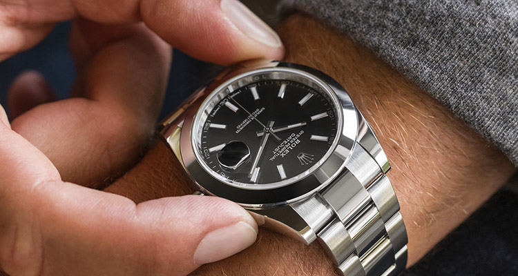 Ein Mann hält seine Rolex Oyster Perpetual Datejust Uhr am Handgelenk zwischen zwei Fingern