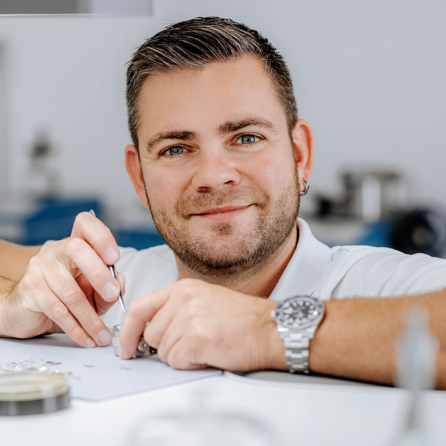 Uhrmachermeister Ronald Förster in Uhrmacherwerkstatt mit Uhrengehäuse und Werkzeug in der Hand.