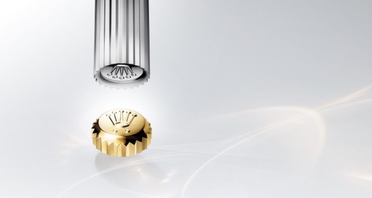 Eine goldene Rolex Aufzugskrone vor hellem Hintergrund mit zwei Punkten unterhalb des Rolex Symbols.